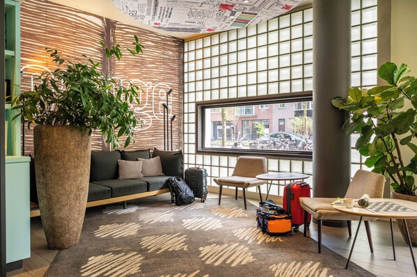 A modern hotel lobby with a large potted plant, a sofa, a small table, and a window with a view of the street. - ibis Amsterdam Centre Stopera (Amsterdam)