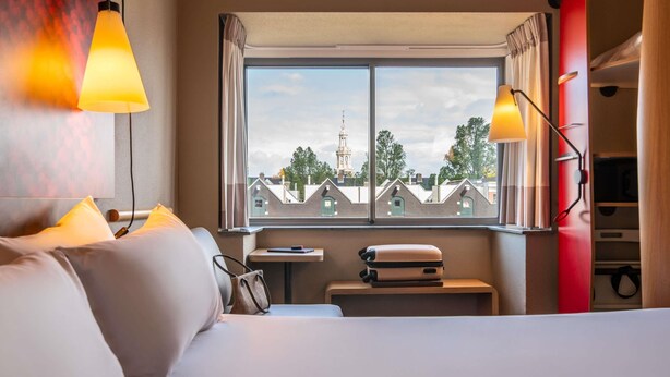 Habitació d'hotel amb un llit, una finestra que mostra edificis, una taula petita i una maleta.