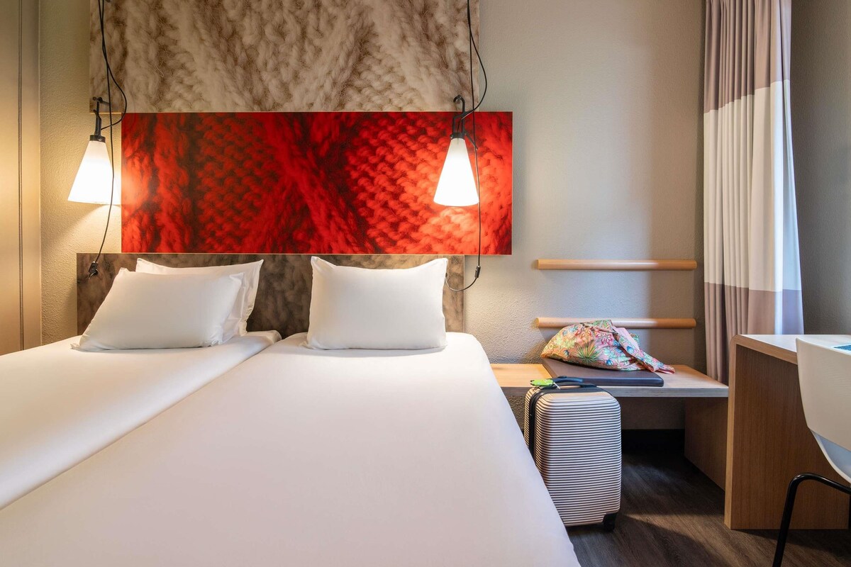A hotel room with two beds, a desk, a chair, a colorful bag, and a red abstract painting.