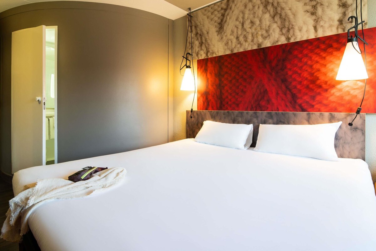 A hotel room with a bed, a headboard with a red and grey patterned design, two wall-mounted lamps, and a door leading to another room.