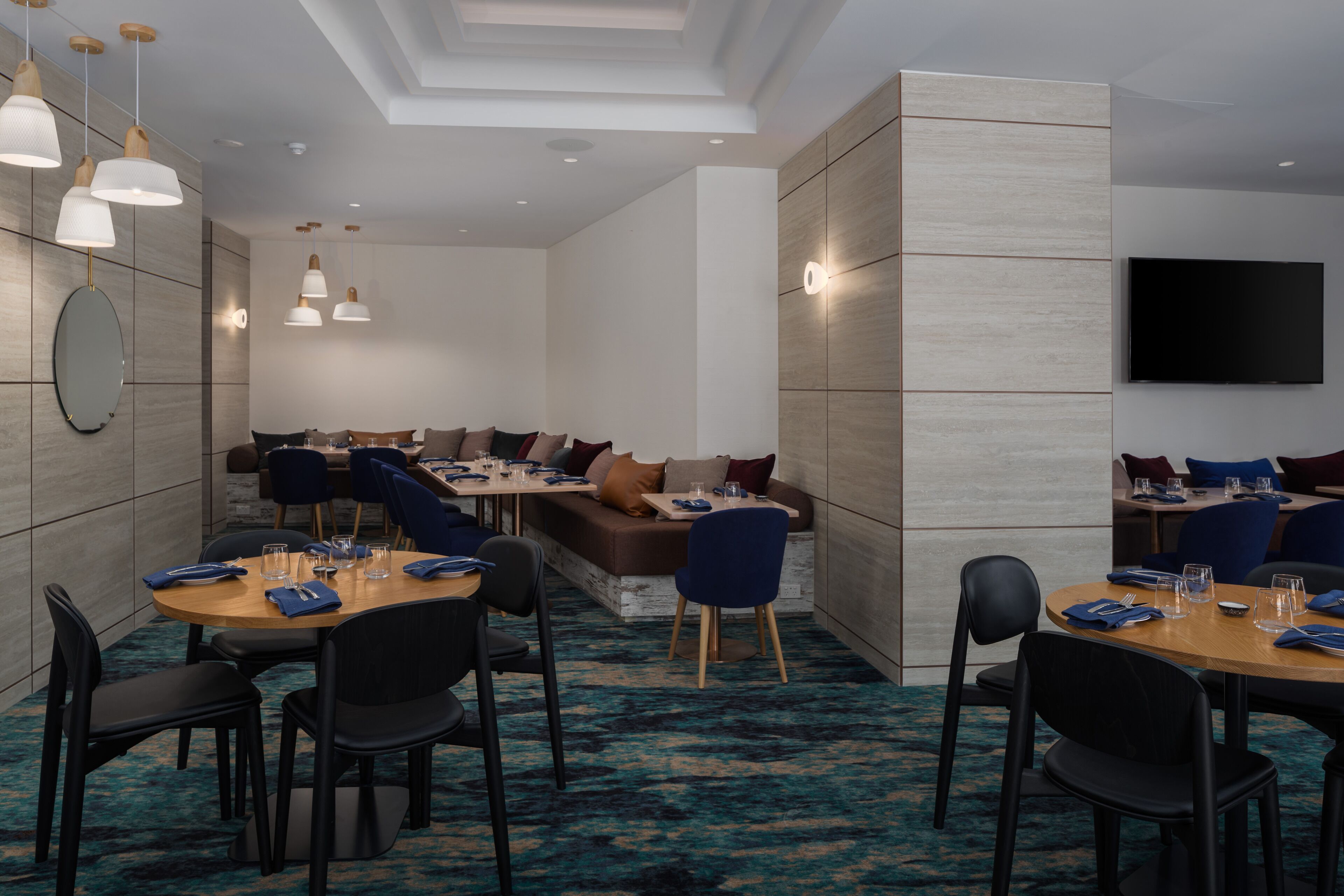 Zona de menjador d'hotel moderna amb taules rodones, cadires blaves i un espai central amb sofà i coixins.