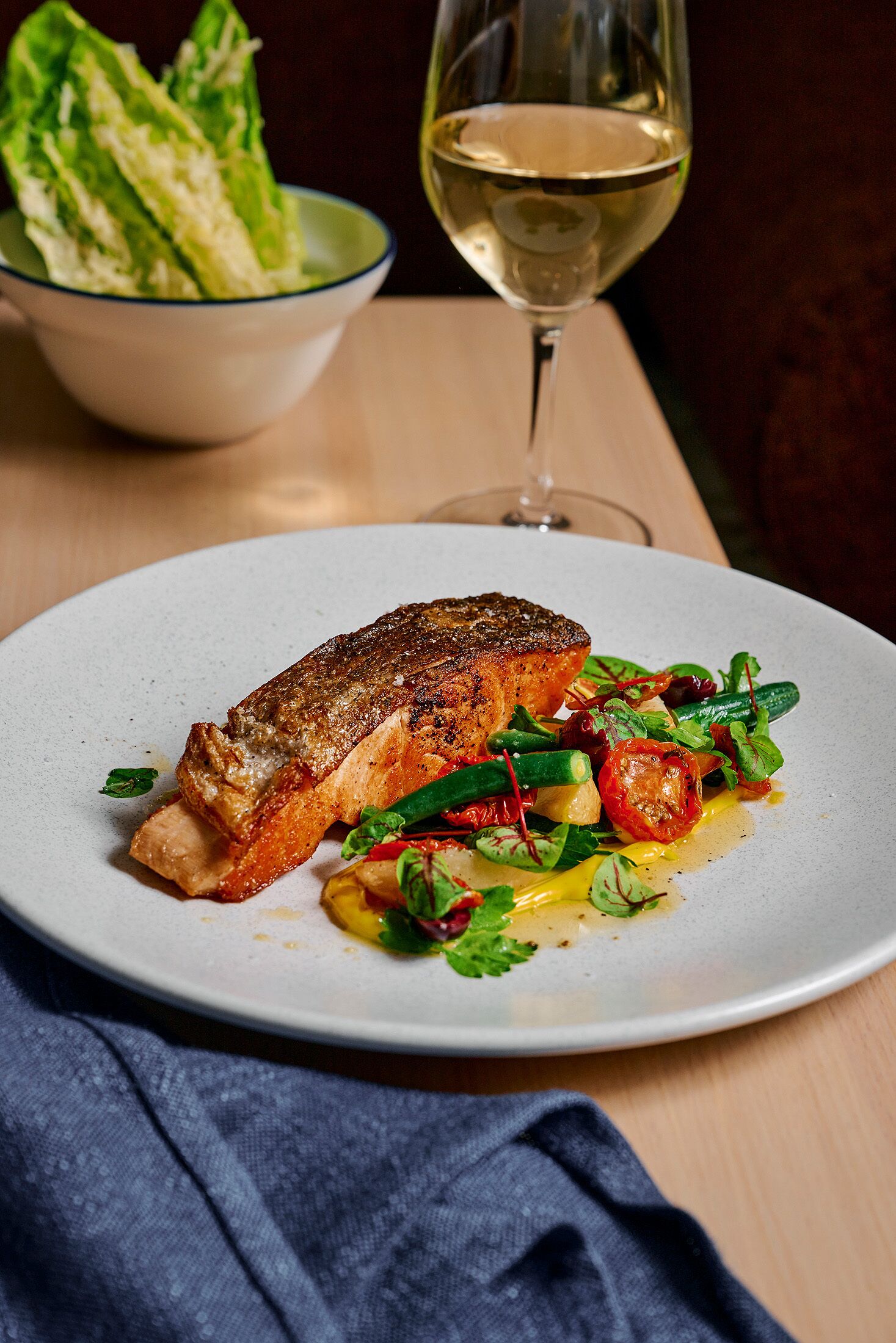 Une assiette de nourriture comprenant un morceau de saumon, des légumes et un verre de vin blanc, le tout posé sur une table en bois.