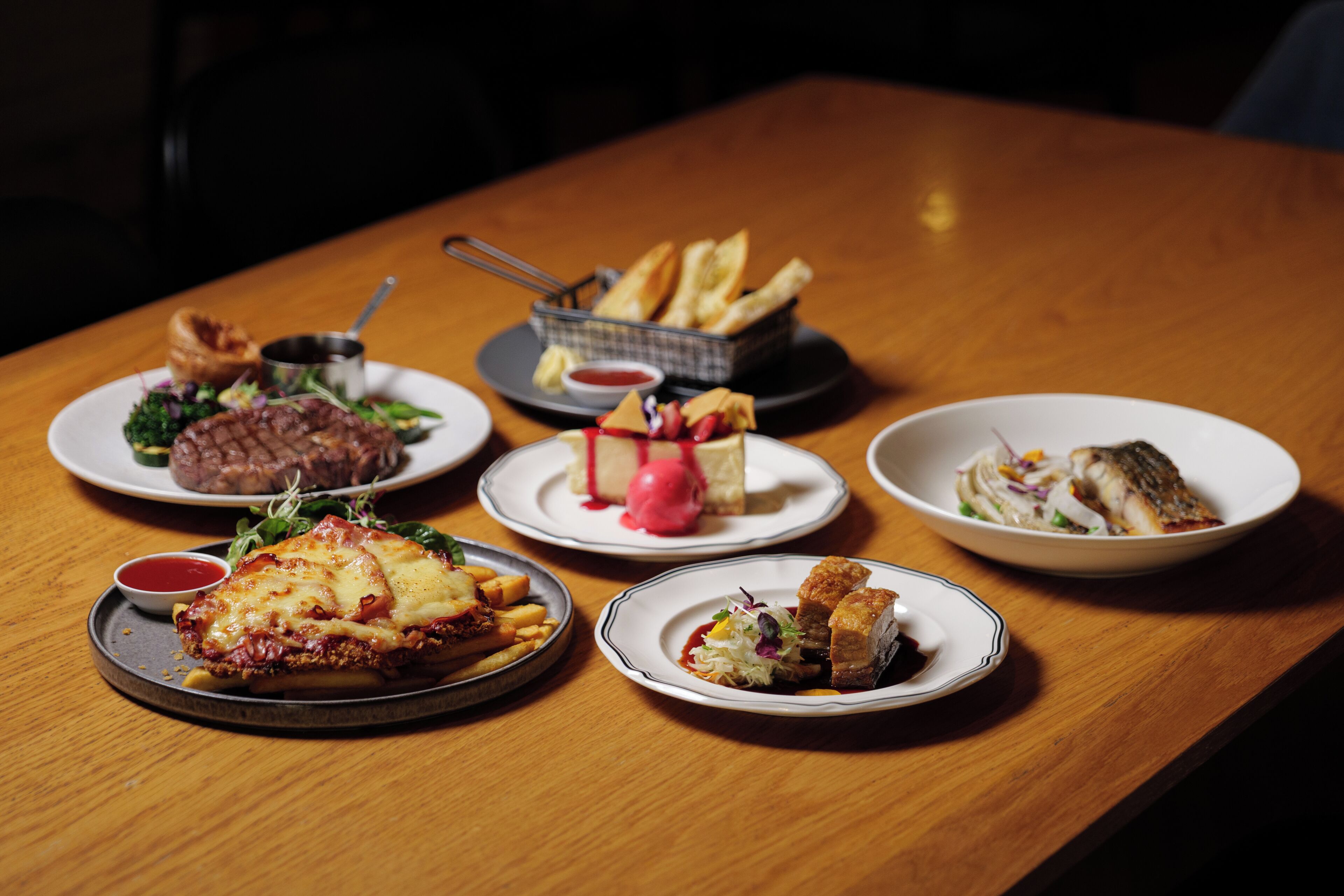 A wooden table with various dishes, including a steak, fries, and a fish dish.