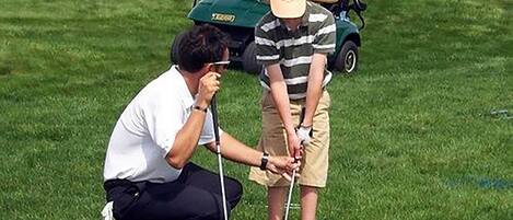 A person in a white shirt and black pants crouching next to another person in a striped shirt and beige shorts, both holding golf clubs on a grassy field with a golf cart in the background.