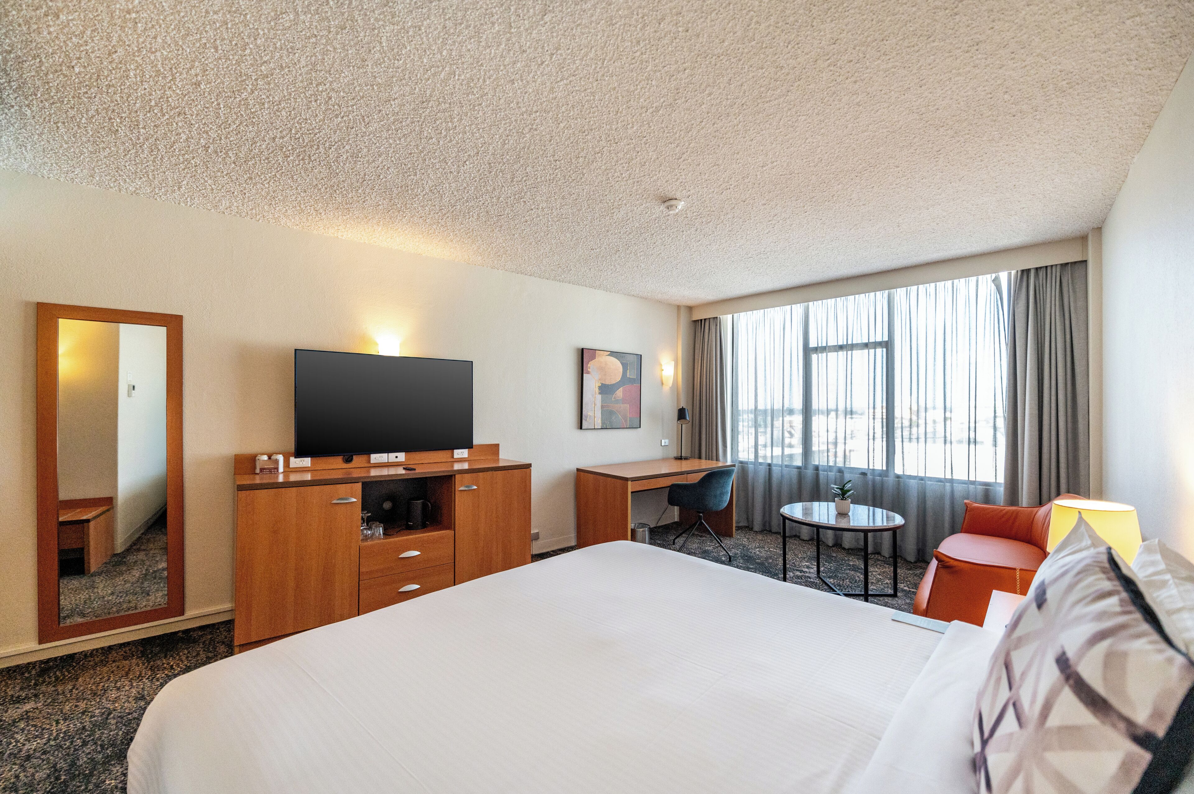 Ett hotellrum med en stor säng, ett skrivbord med en platt-TV, ett litet bord med en stol, en spegel och ett fönster med gardiner.