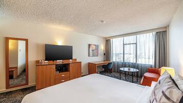 A hotel room with a large bed, a desk with a flat-screen TV, a small table with a chair, a mirror, and a window with curtains.