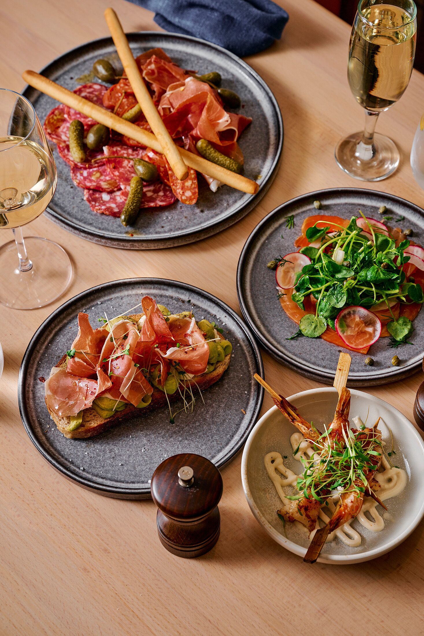 A table with various dishes, including a plate with sliced meats and olives, a plate with a salad, and a bowl with a skewer of food. There are also glasses of wine and a pepper grinder on the table.