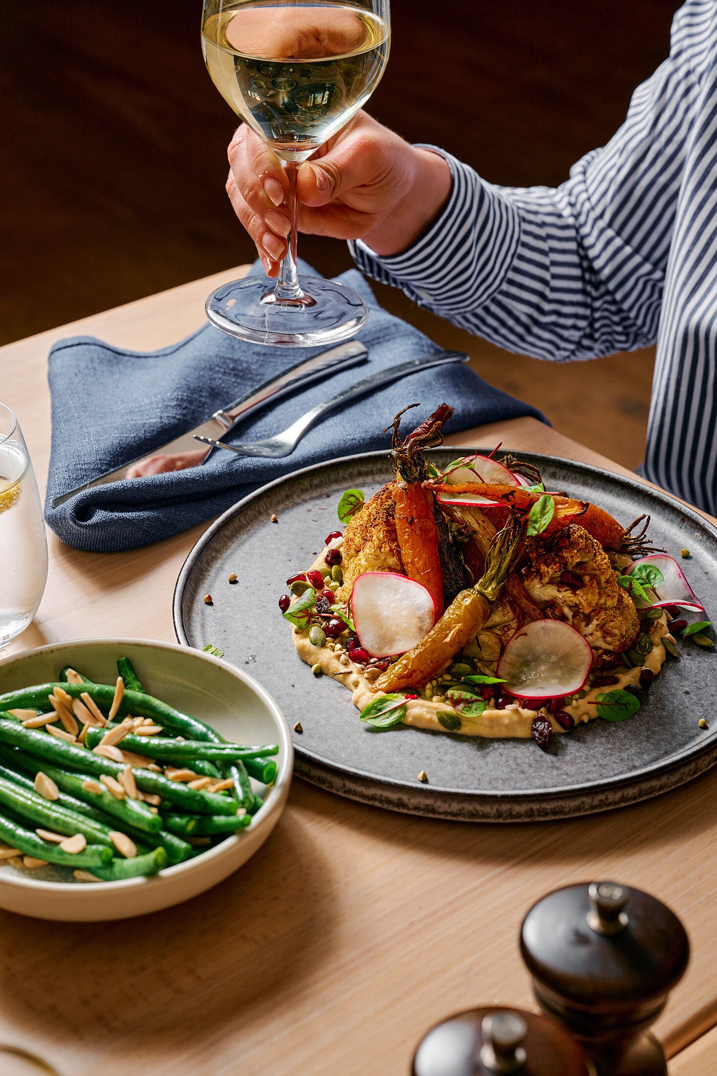 A dining table with a plate of food, a glass of white wine, and a bowl of green beans.