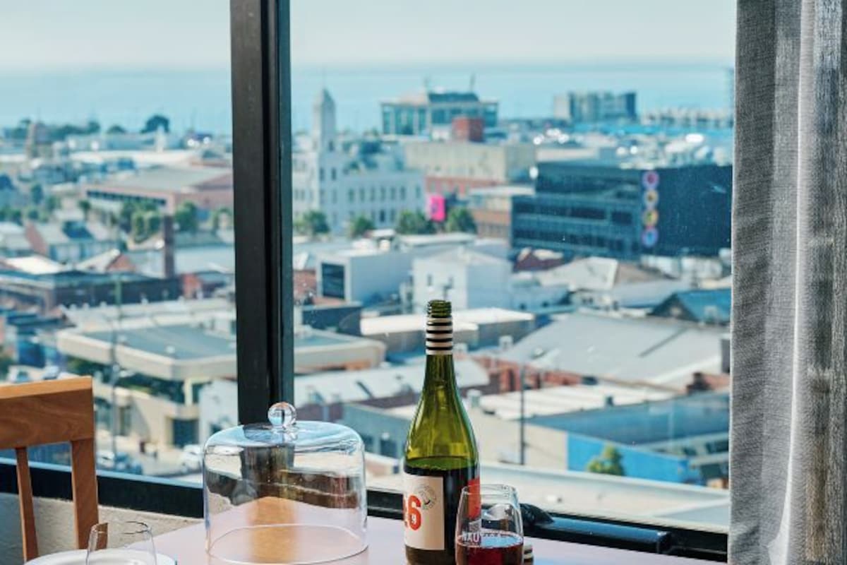 Una mesa con una botella de vino, una copa y un recipiente de vidrio transparente, con vistas a un paisaje urbano a través de una ventana.