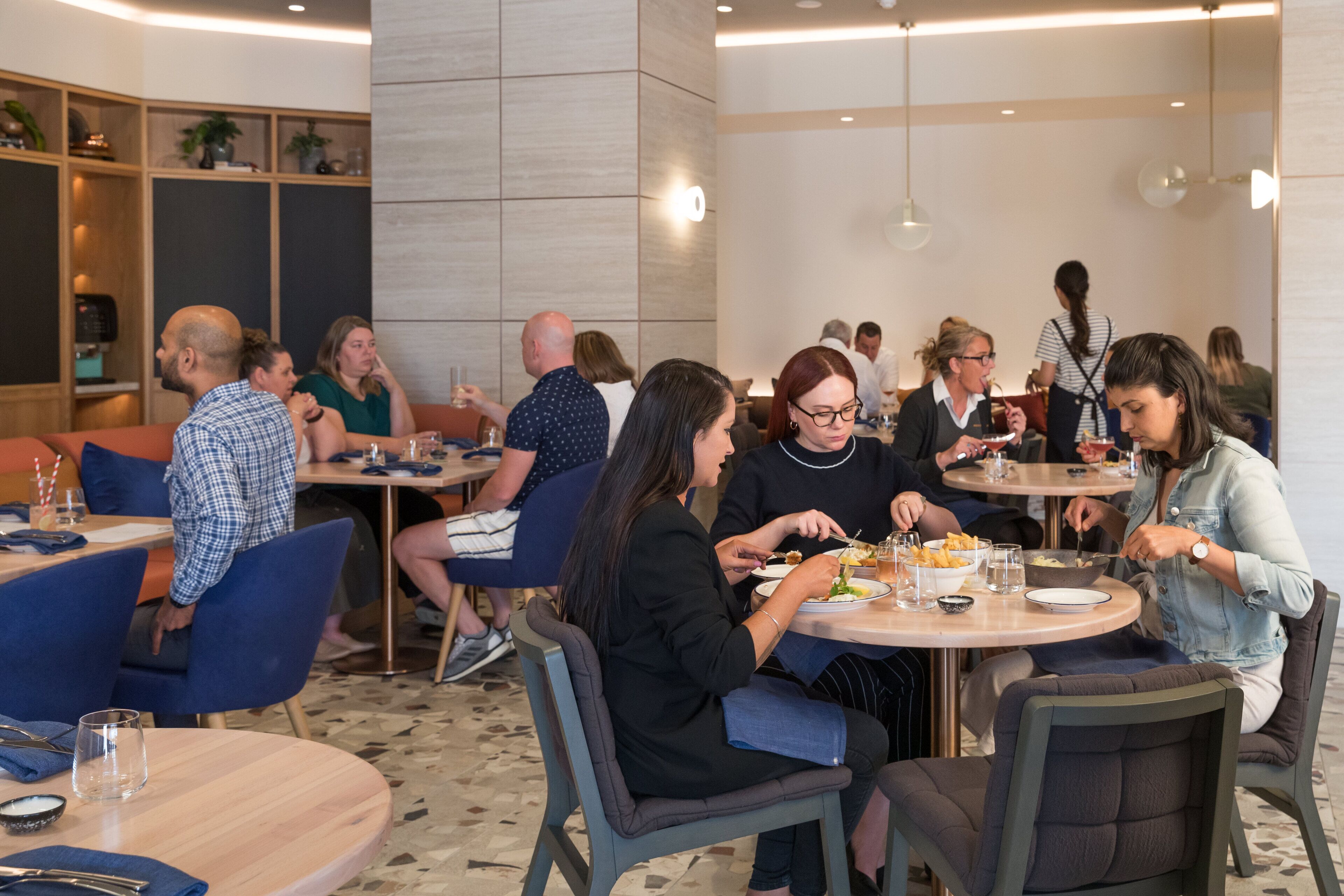 A modern dining area with round tables and blue chairs, featuring a clean, contemporary design with neutral colors and a mix of seating arrangements.