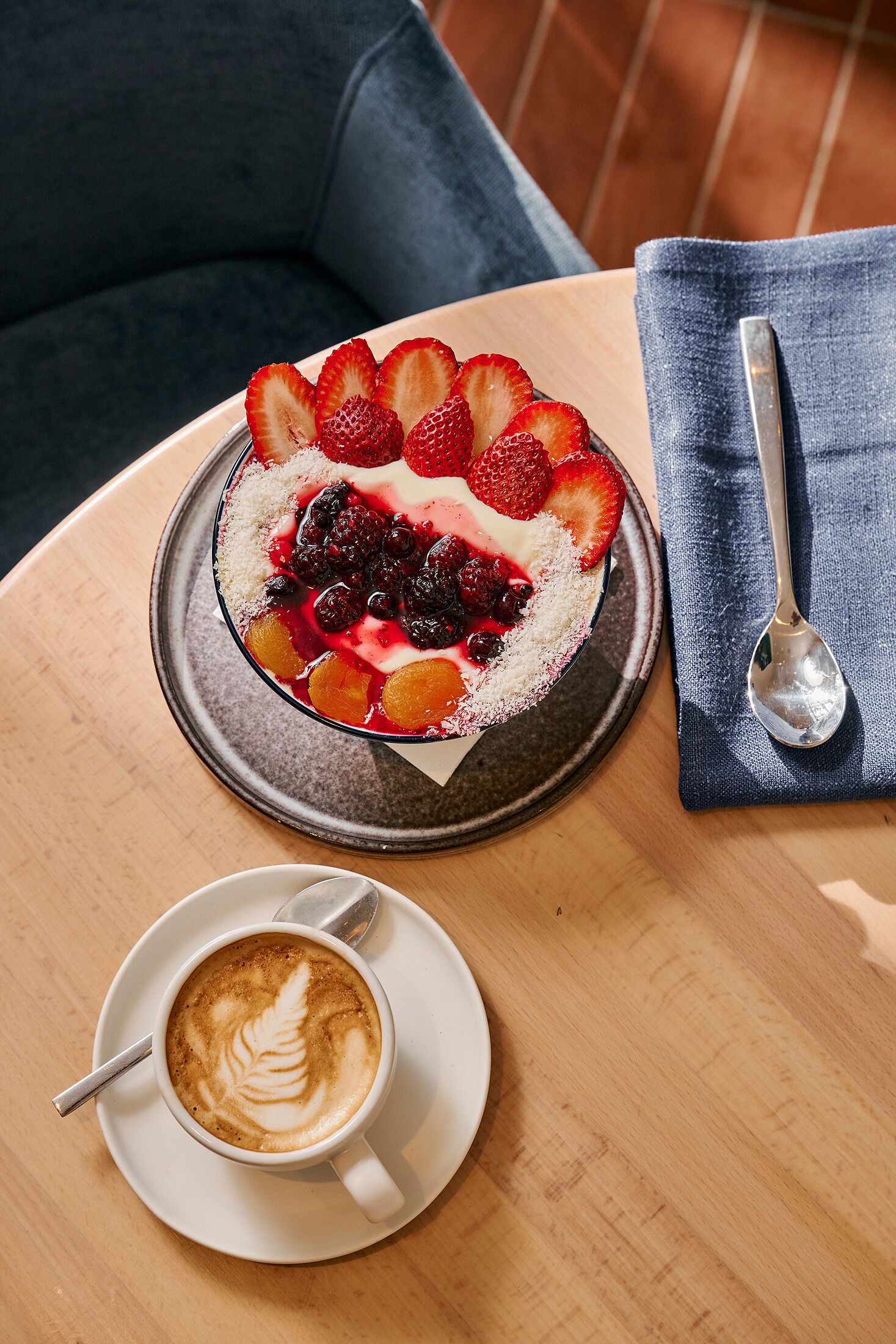 A dessert plate with strawberries and berries, a cup of coffee with latte art, and a spoon on a napkin on a wooden table.