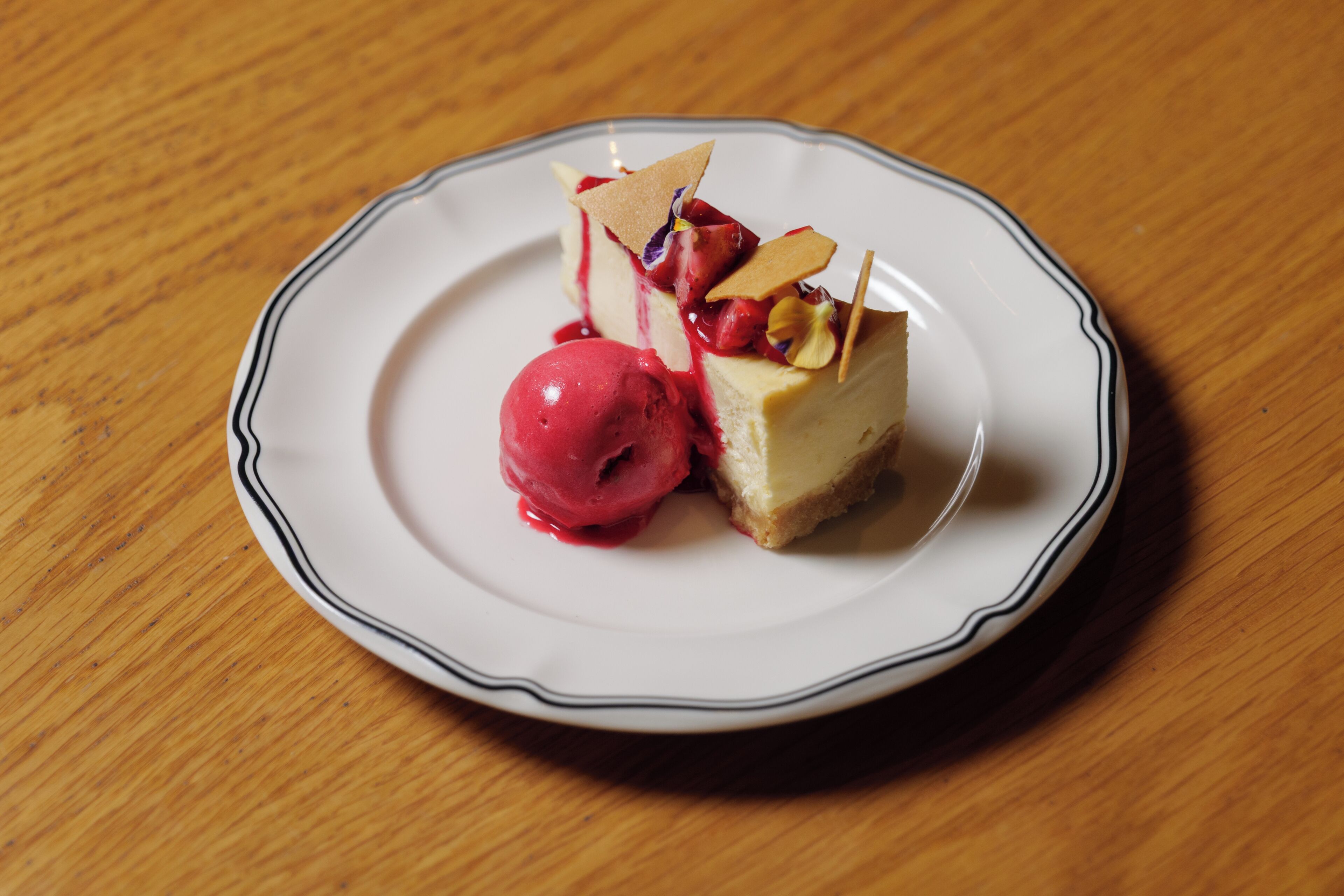 Sobremesa em um prato branco com borda preta, com uma fatia de cheesecake, uma bola de sorvete vermelho e um purê de frutas vermelhas.