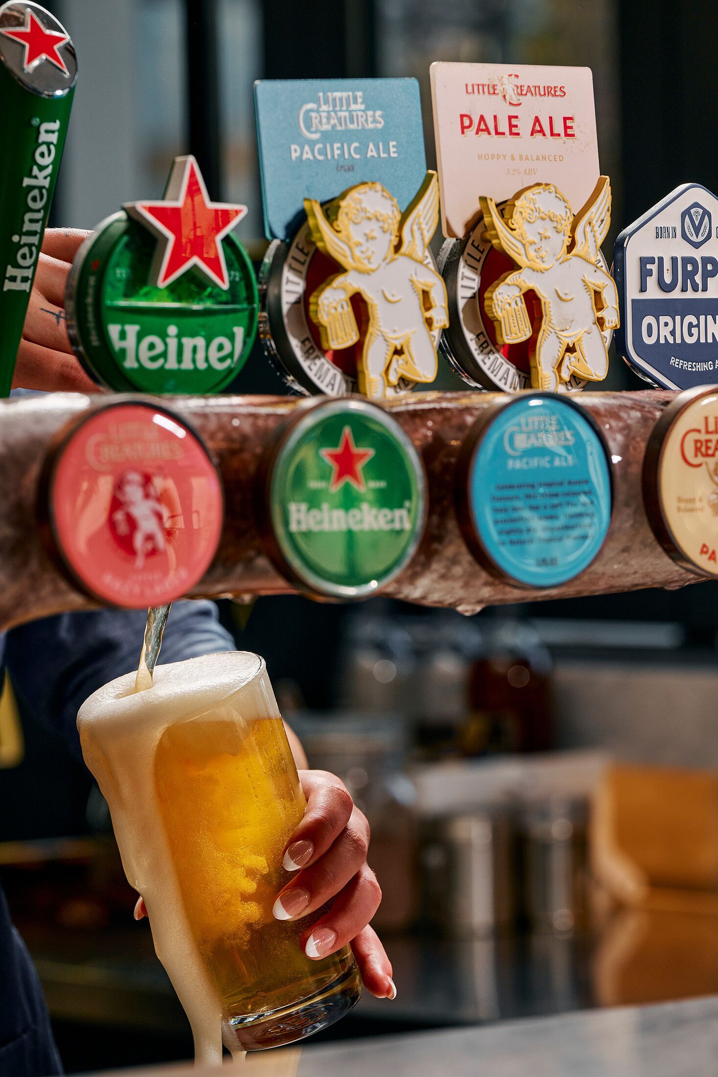 A hand holding a glass of beer with a tap in the background displaying various beer brands.