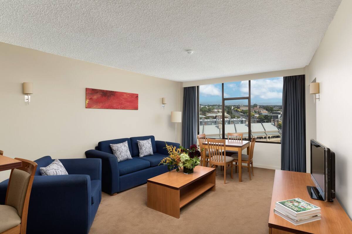 Una habitación de hotel moderna con un sofá azul, una mesa de centro de madera, una mesa de comedor con sillas y un ventanal con vistas a la ciudad.