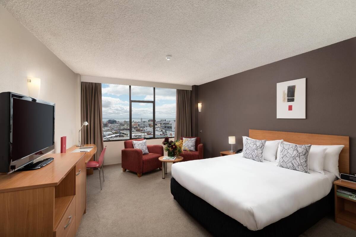 Habitación de hotel con una cama grande, un escritorio con televisor, dos sillones, una mesita con un jarrón y vistas a la ciudad a través de amplias ventanas.