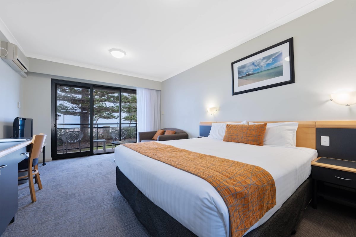 A hotel room with a large bed, a desk, a chair, a TV, and a balcony with a view of trees and a building.