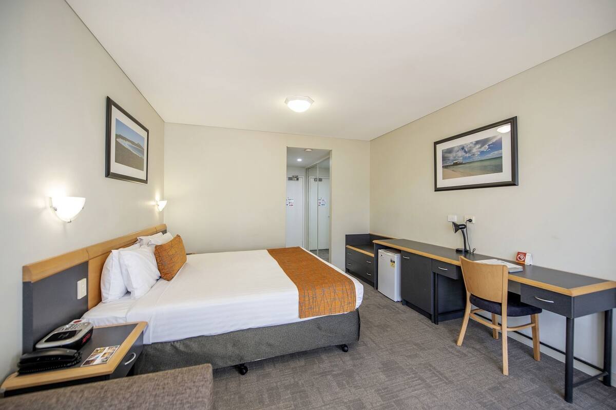 A hotel room with a bed, desk, chair, and a small refrigerator.