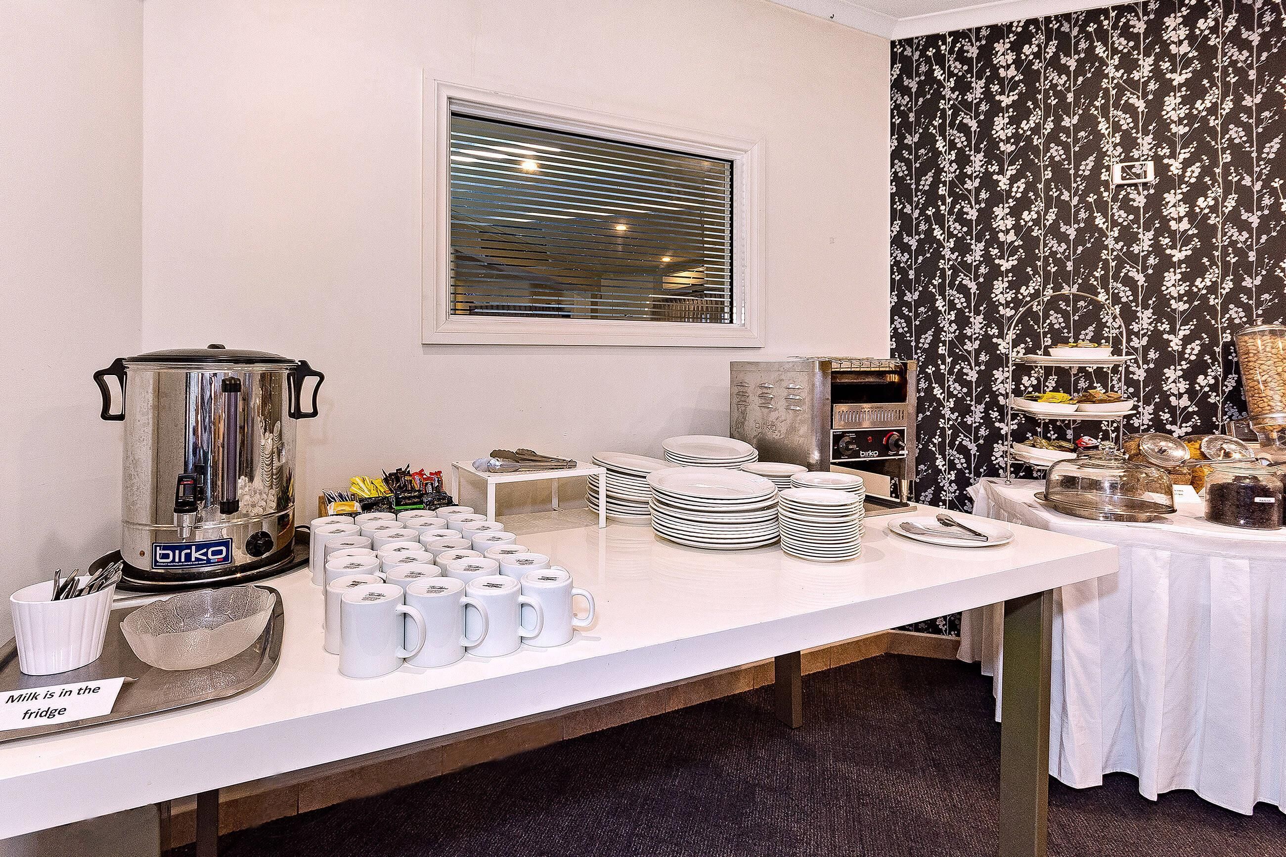 Disposición de bufé con cafetera, tazas, platos y refrigerador.