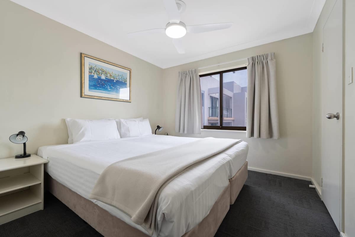 A hotel room with a bed, a desk, a ceiling fan, a window with curtains, and a painting on the wall.