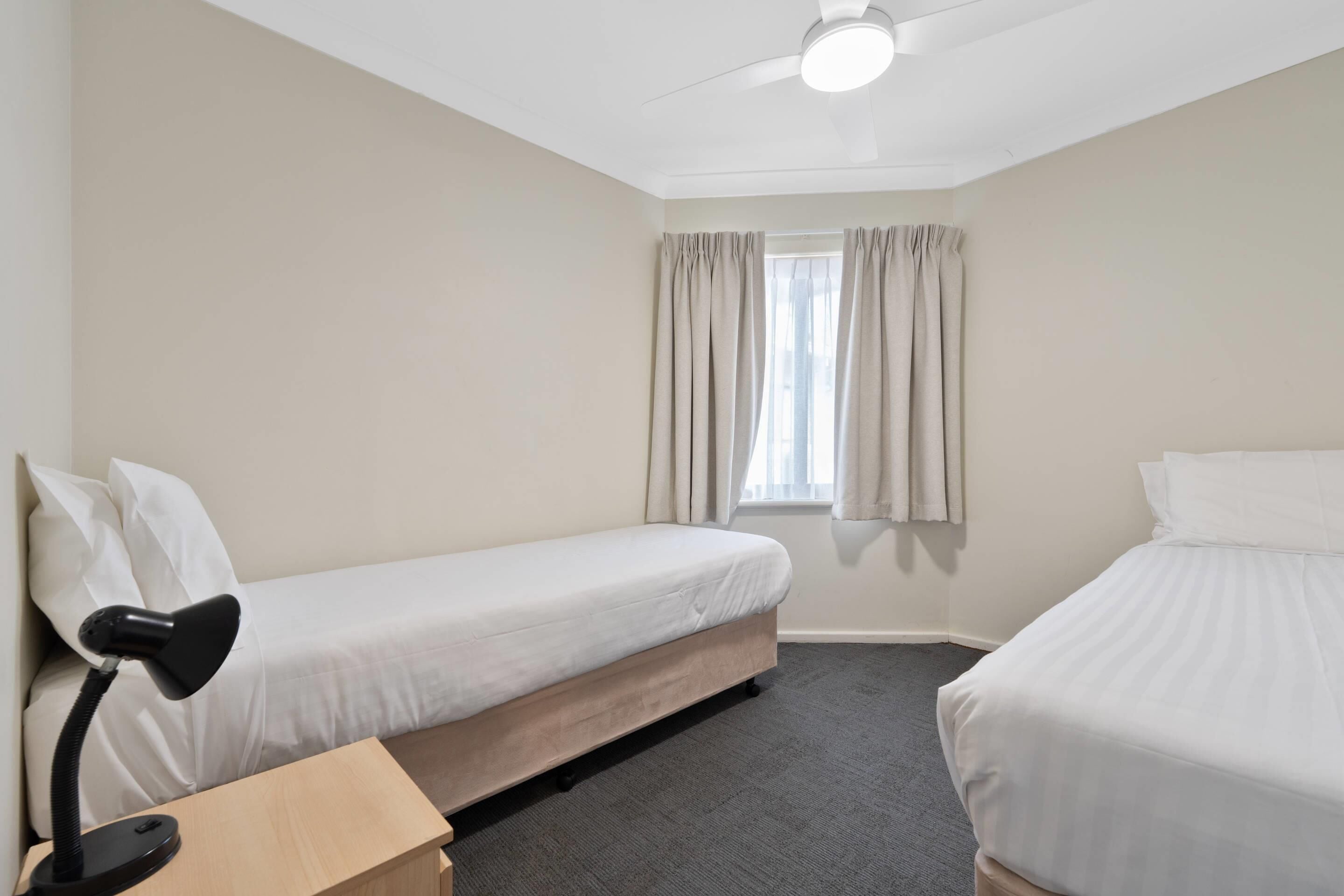 Ett hotellrum med två separata sängar, ett skrivbord med en lampa, en takfläkt och ett fönster med gardiner.