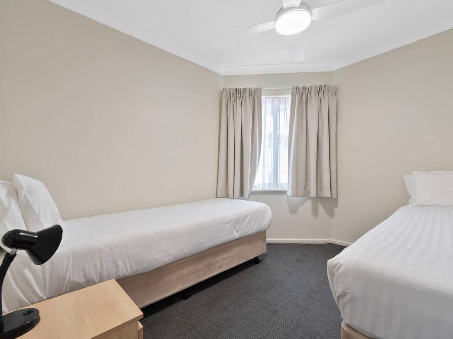 Ett hotellrum med två separata sängar, ett skrivbord med en lampa, en takfläkt och ett fönster med gardiner.