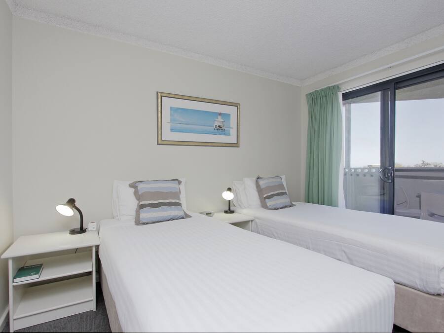 Habitación de hotel con dos camas, una mesita de noche con lámpara, un cuadro en la pared y una ventana con vistas.