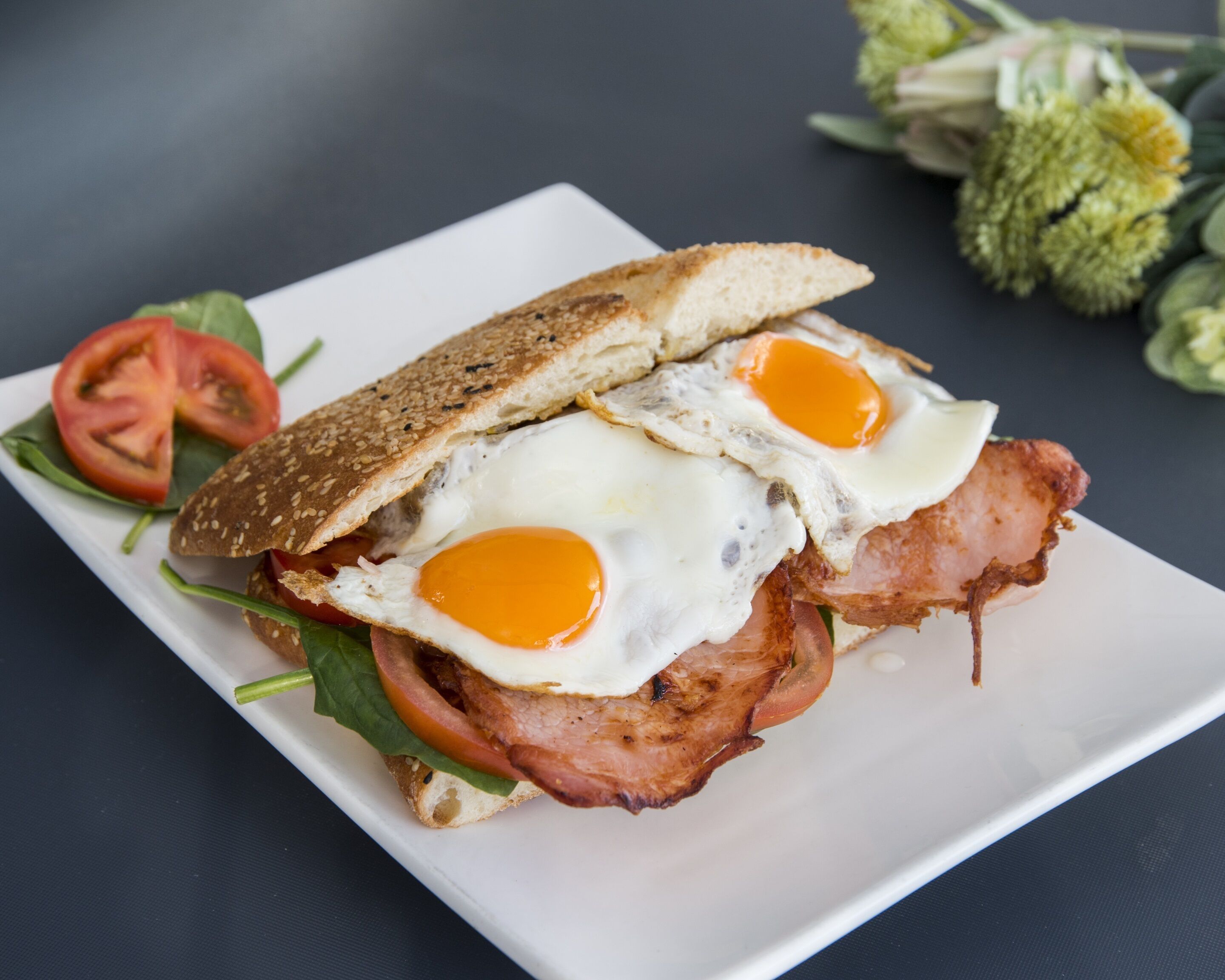 Un sandwich aux œufs et au bacon, servi sur une assiette blanche, accompagné d’une tranche de tomate et de quelques feuilles de salade.