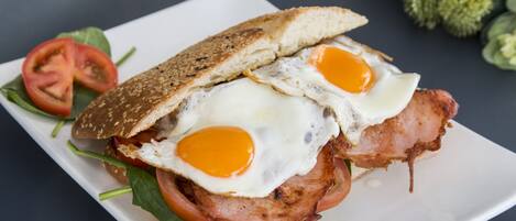 Un sandwich aux œufs et au bacon, servi sur une assiette blanche, accompagné d’une tranche de tomate et de quelques feuilles de salade.