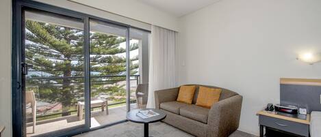 A hotel room with a sofa, a small table, a chair, a nightstand, and a large sliding glass door leading to an outdoor area with a view of trees and a fence.