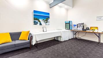 A conference room with a white table, a sofa with yellow cushions, a painting of a beach, and a water dispenser.