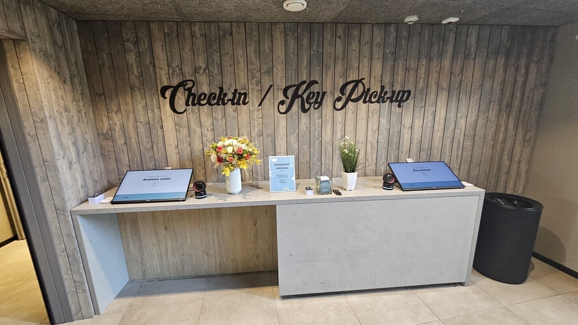 A reception desk with two laptops, a sign for key pickup, and a small potted plant.