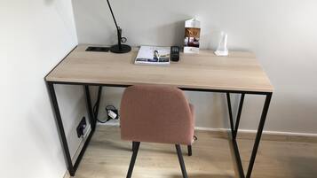 En arbetsplats med ett skrivbord i trä, en svart skrivbordslampa, en rosa stol, en fjärrkontroll, en tidning, en mjölkförpackning och en plastmugg.