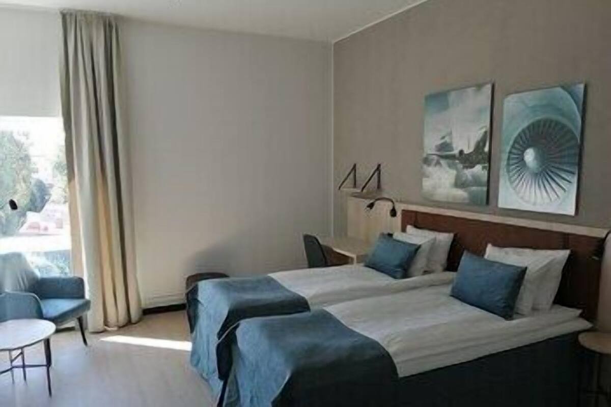 A hotel room with two beds, a small table, a chair, and a window with curtains.