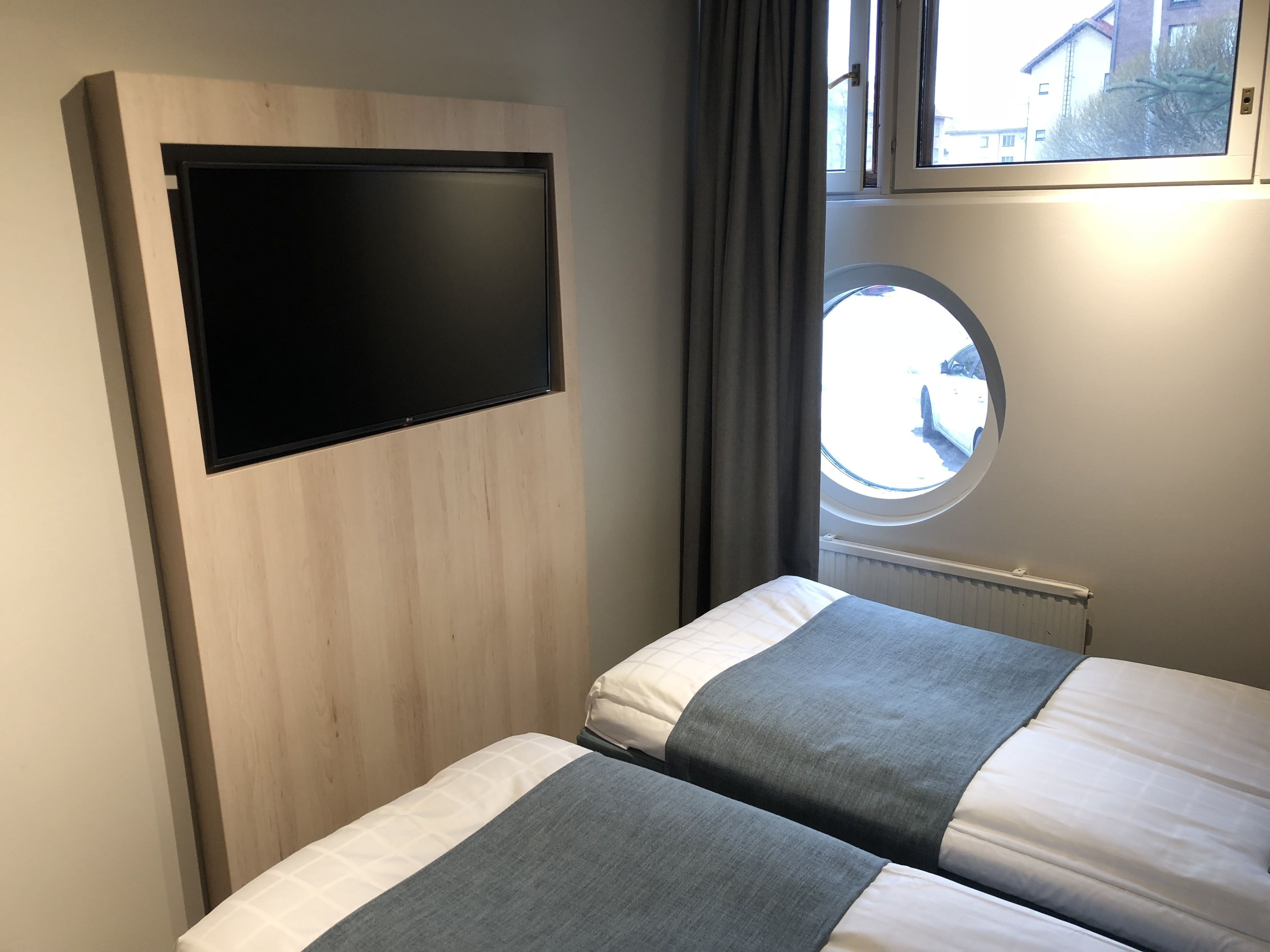 Un dormitorio con dos camas, un televisor montado en un panel de madera, una ventana con vistas a edificios y un radiador.