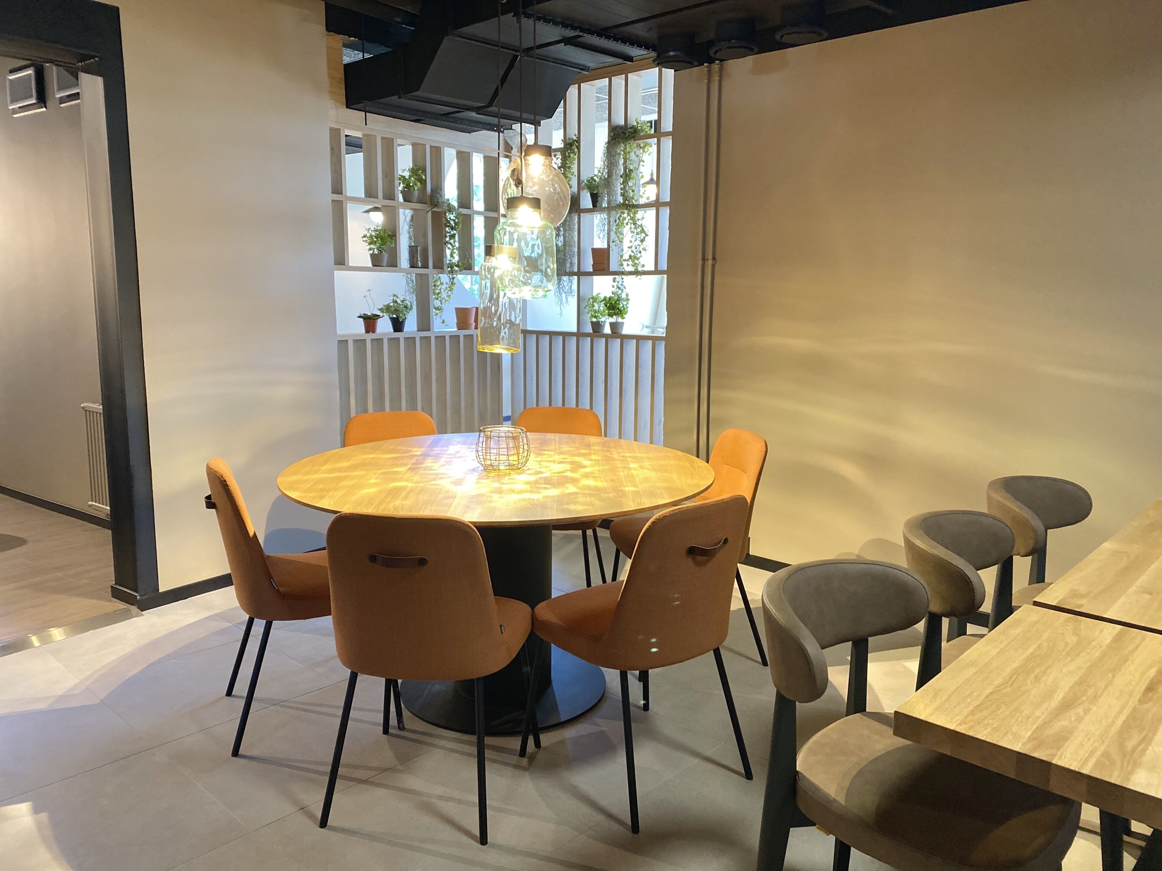 A modern meeting room with a round wooden table surrounded by orange chairs, a long table with grey chairs, and a white partition with potted plants.