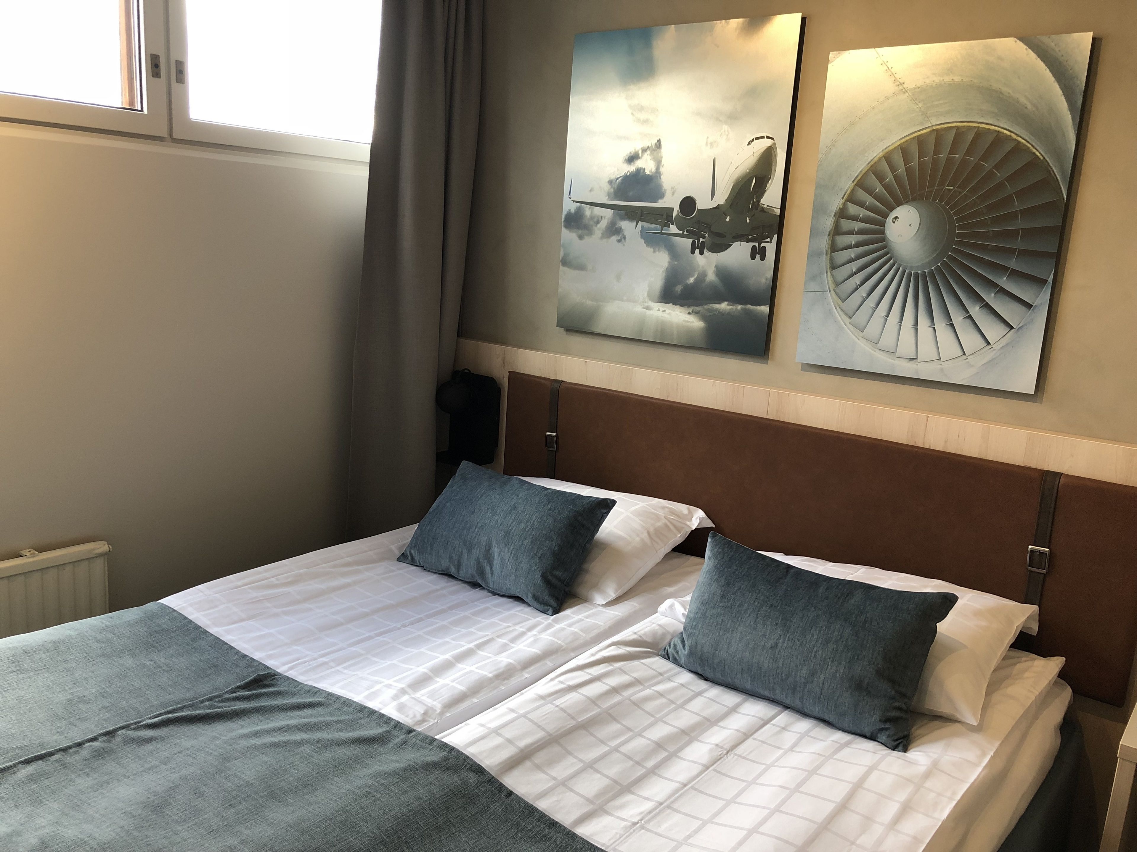 Quarto de hotel com cama, duas almofadas, janela com cortinas e duas obras de arte na parede retratando um avião e um ventilador.