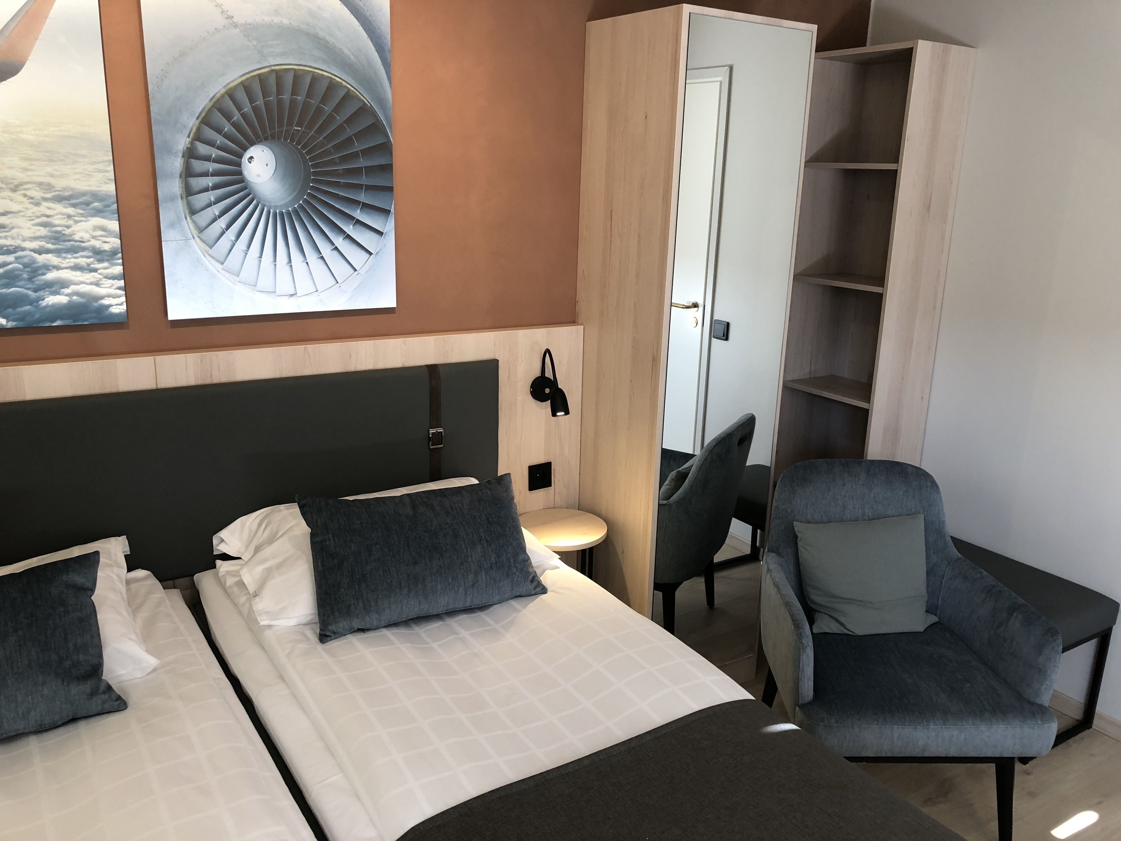 A hotel room with a bed, two chairs, a desk, a wardrobe, and a large window with a view of clouds and an airplane engine.