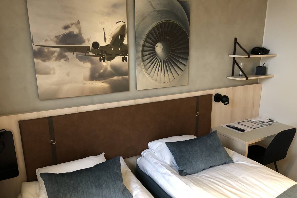 A hotel room with two beds, a desk, and a chair. There are wall art pieces depicting an airplane and a jet engine.