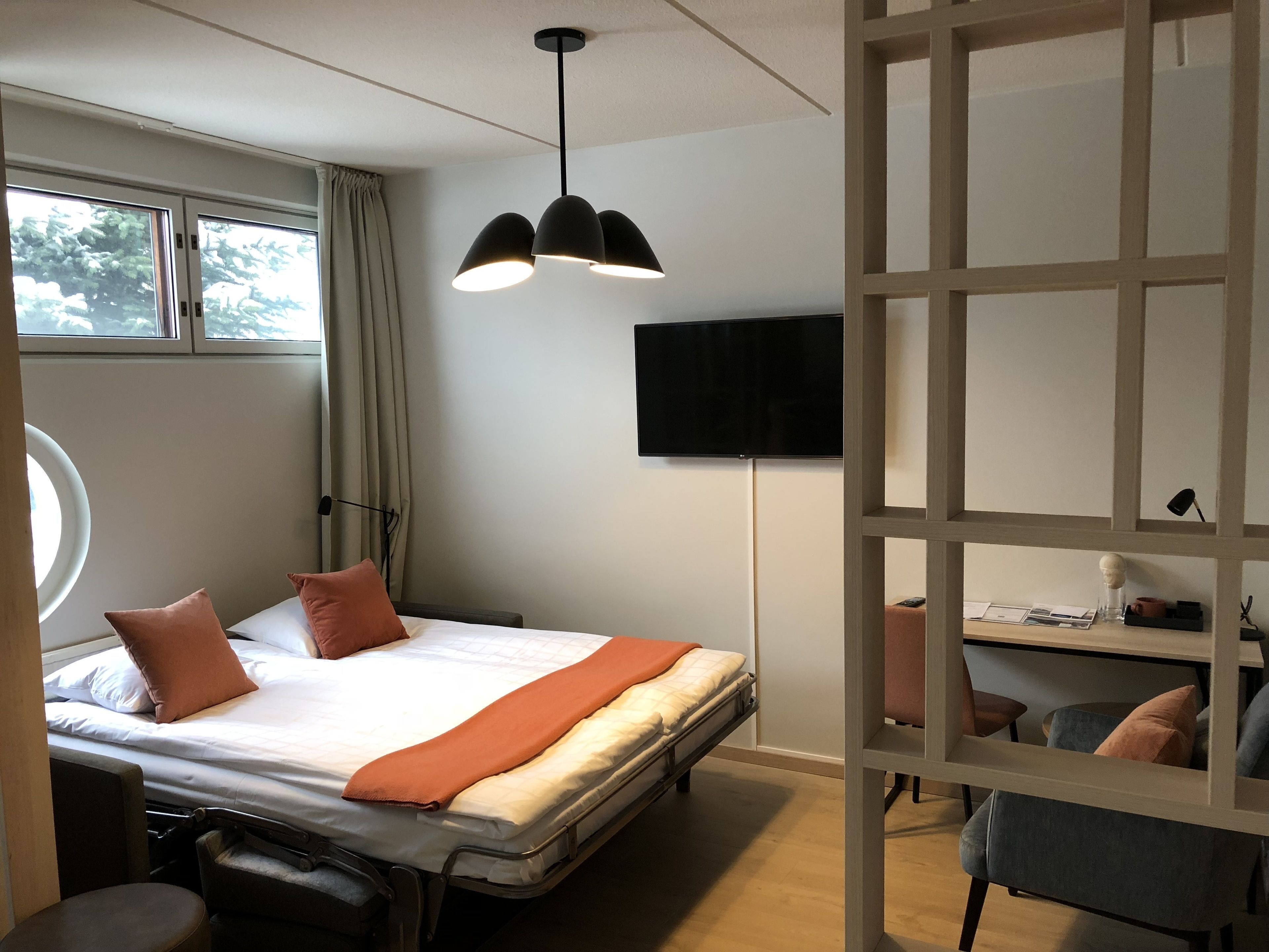 Habitación de hotel con cama, escritorio, silla, televisión y ventana con cortinas.