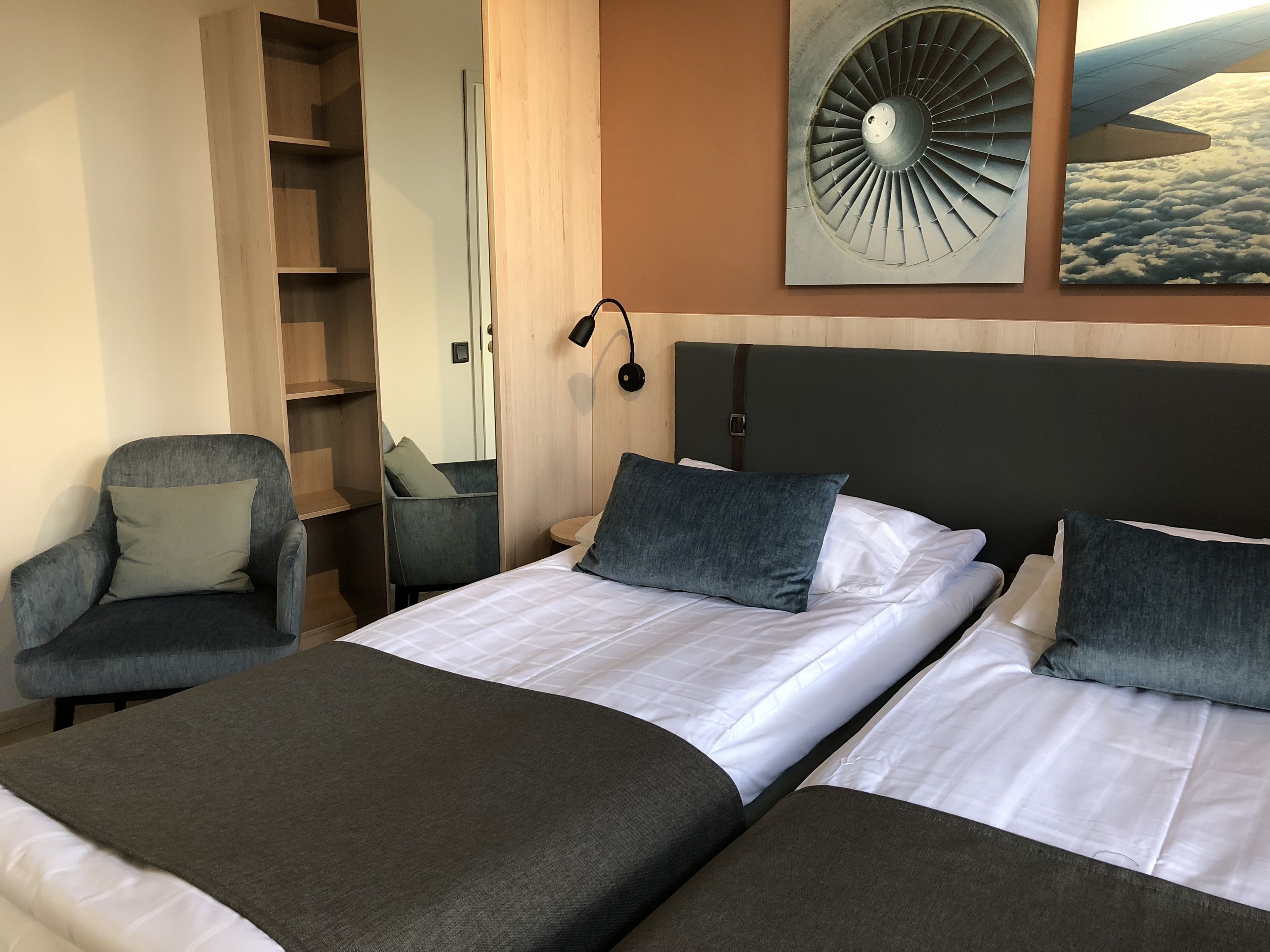 Quarto de hotel com duas camas, uma cadeira, um guarda-roupa e um ventilador fixado na parede.