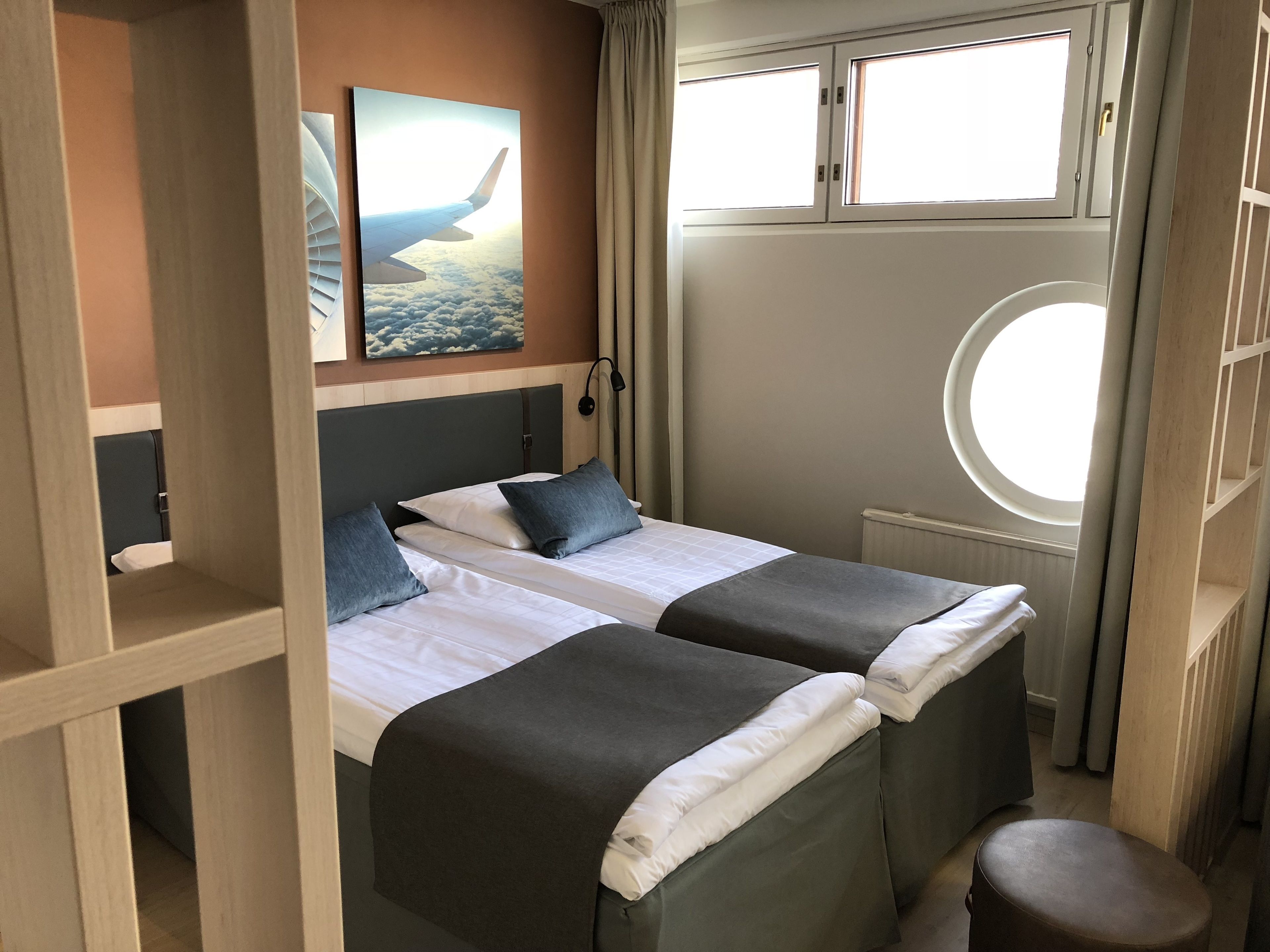 A hotel room with two beds, a window with curtains, a wall-mounted picture, and a small stool.