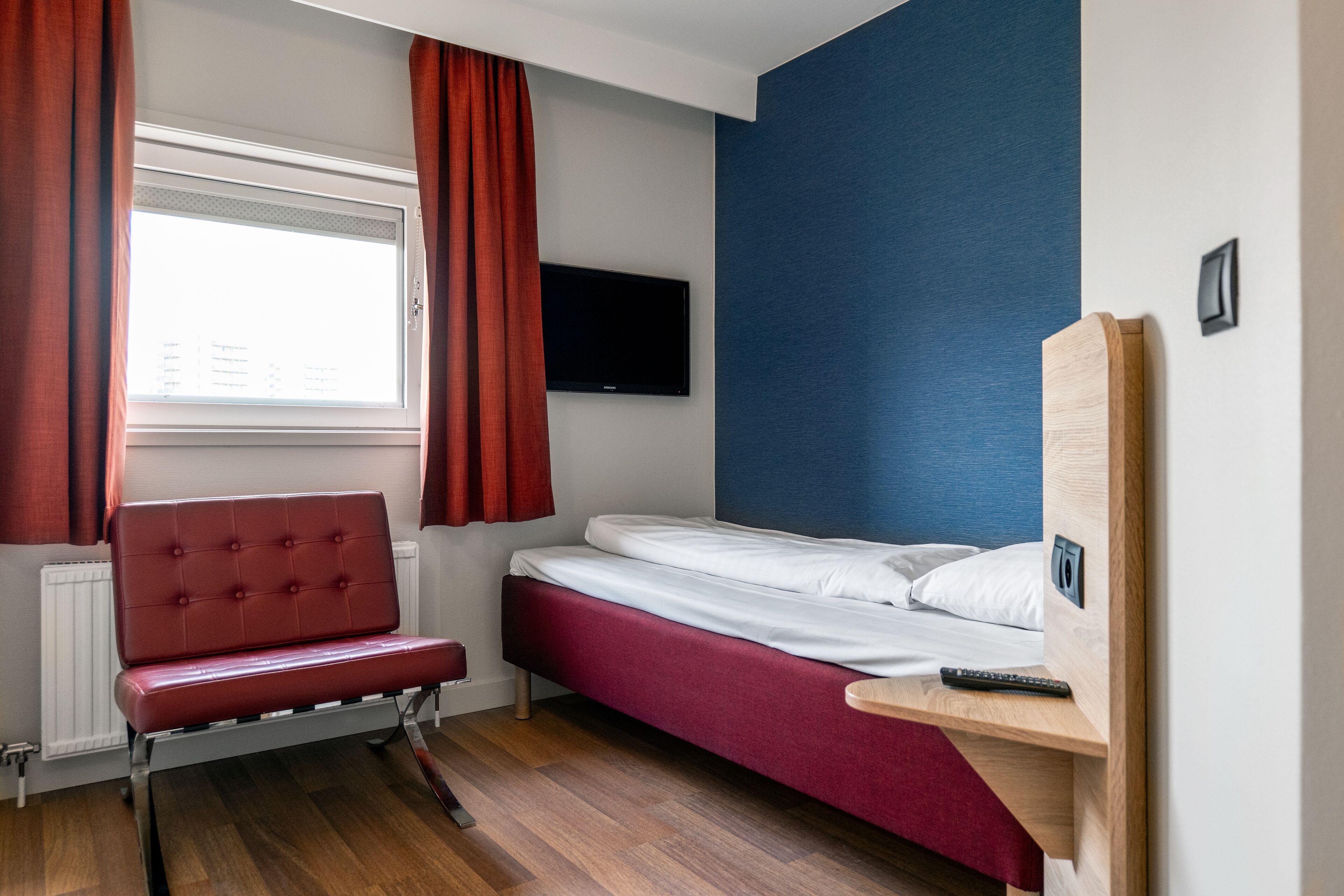Habitación de hotel pequeña con una cama individual, una butaca tapizada en rojo, un televisor montado en la pared y una ventana con cortinas rojas.