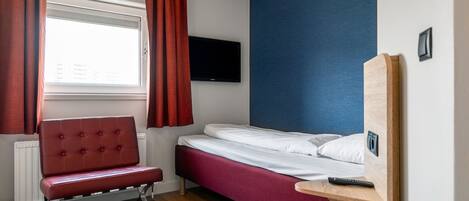 Habitación de hotel pequeña con una cama individual, una butaca tapizada en rojo, un televisor montado en la pared y una ventana con cortinas rojas.