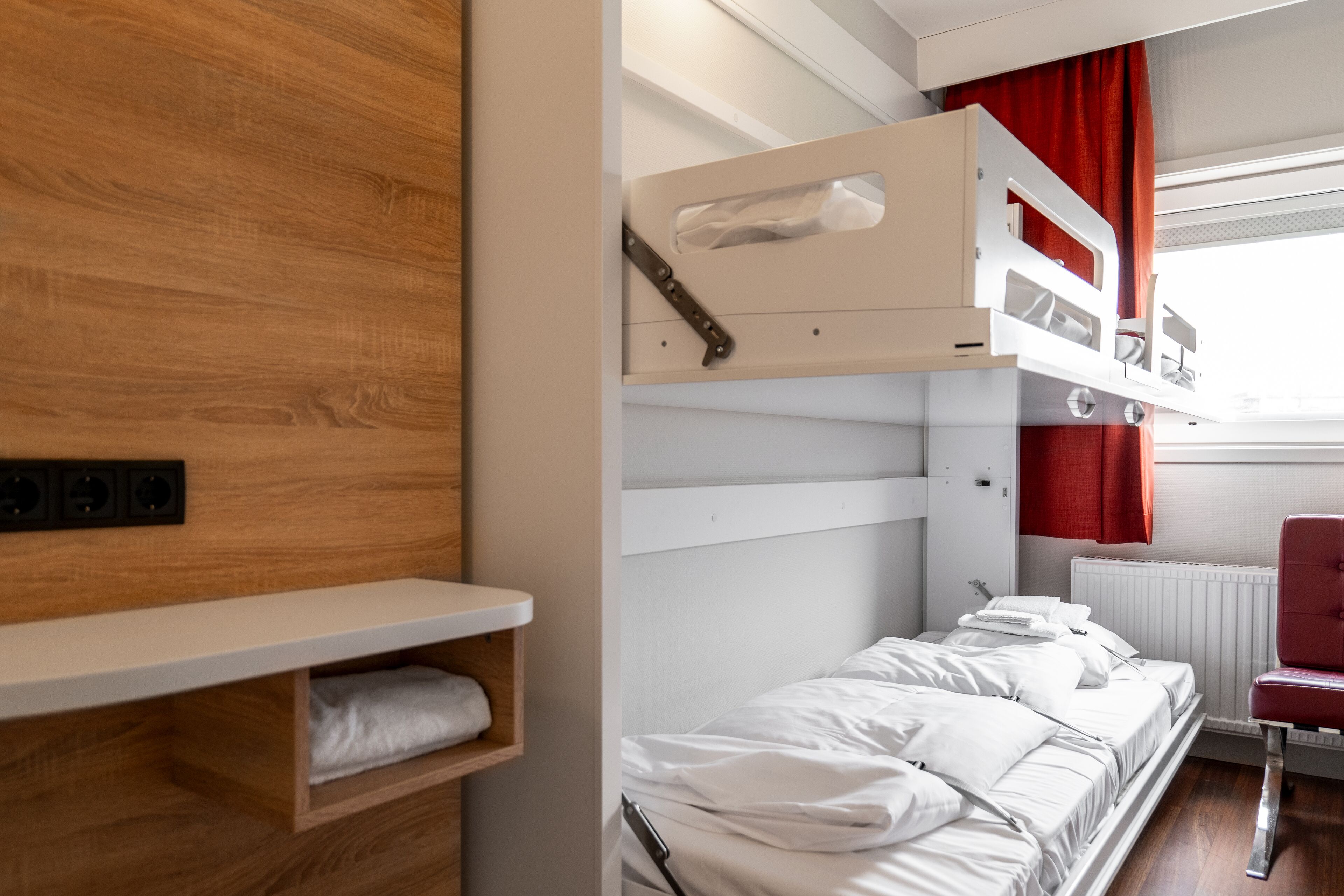Una cama litera con un estante de madera abajo, una silla roja y un radiador blanco.
