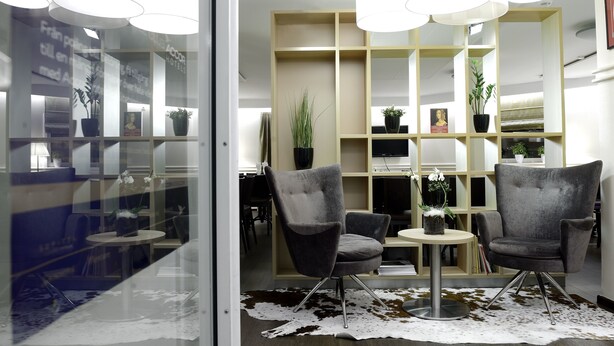 A modern hotel lobby with a glass partition, two grey armchairs, a round table, and a wooden bookshelf with plants. - Good Morning Mölndal (Mölndal)