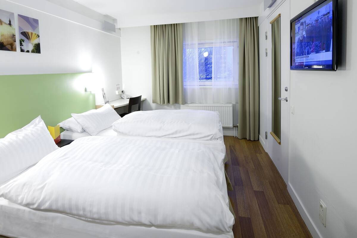 Ett hotellrum med en stor säng, ett skrivbord, en stol, en tv och ett fönster med gardiner.