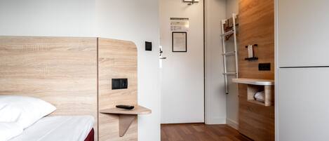 Habitación de hotel compacta con cabecera de madera, mesita de noche pequeña, estante empotrado, cama blanca, suelo de madera, armario blanco y puerta que da a un baño.
