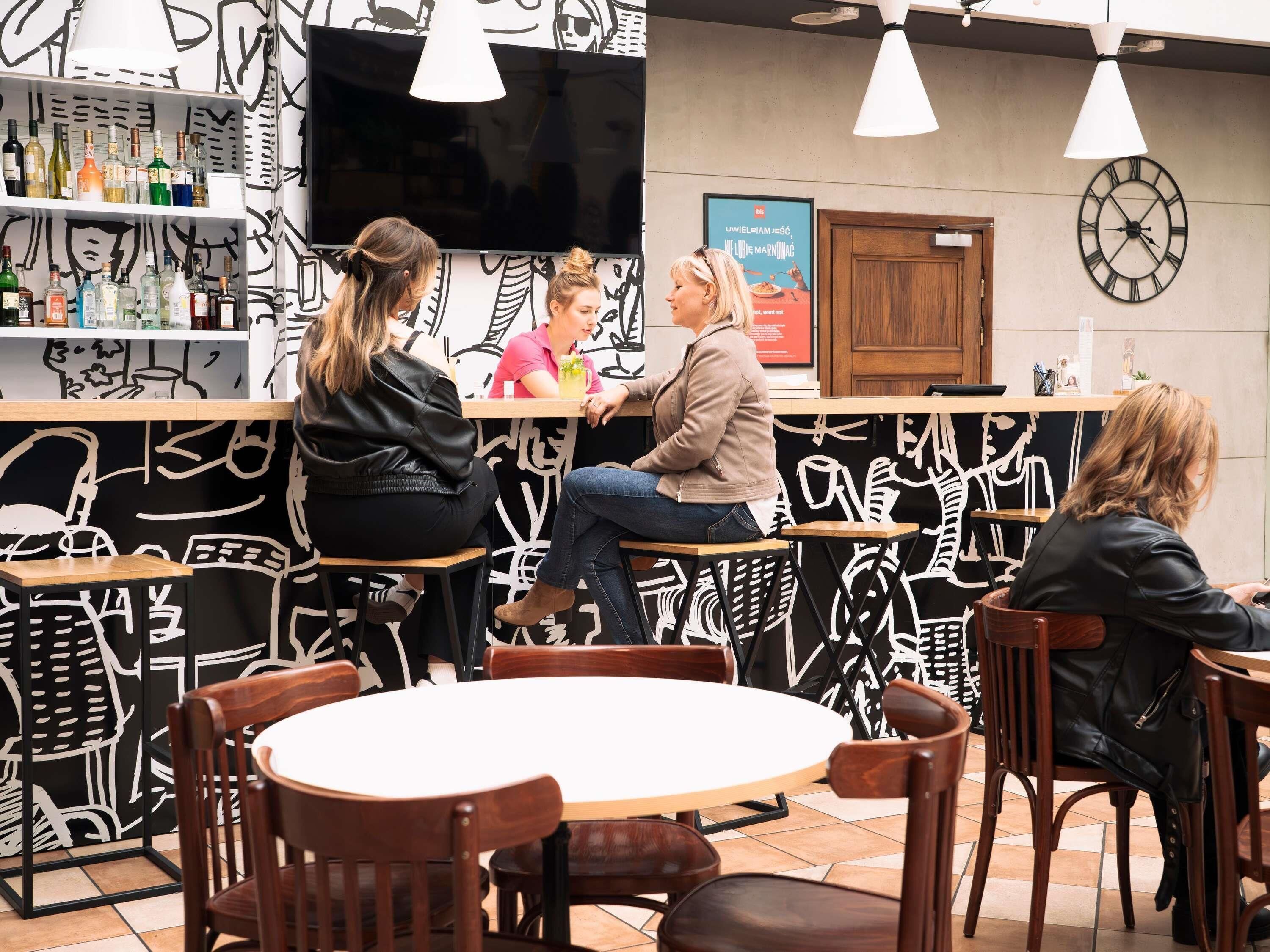Un café con mesas y sillas de madera, una zona de bar con botellas y un reloj de pared.