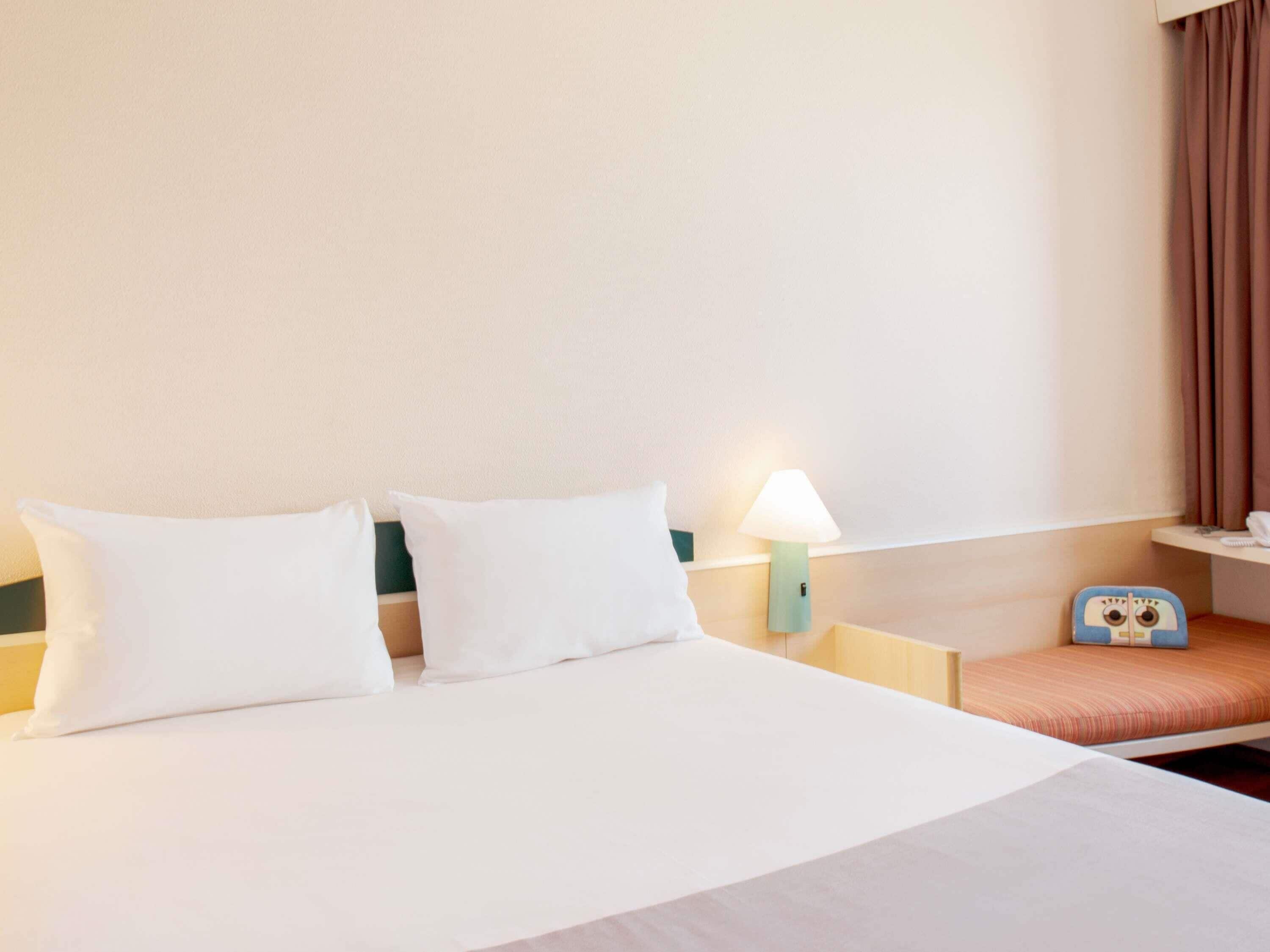 A hotel room with a bed, a nightstand with a lamp, a small table with a telephone, and a window with curtains.