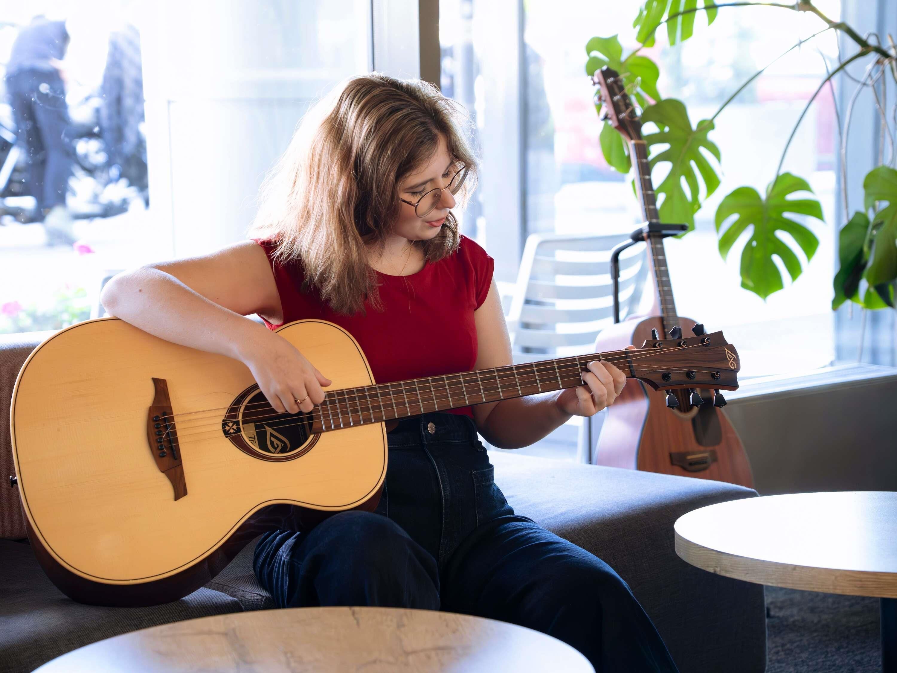 Una persona tocando una guitarra acústica en una habitación con un ventanal grande, una planta y una mesa redonda.